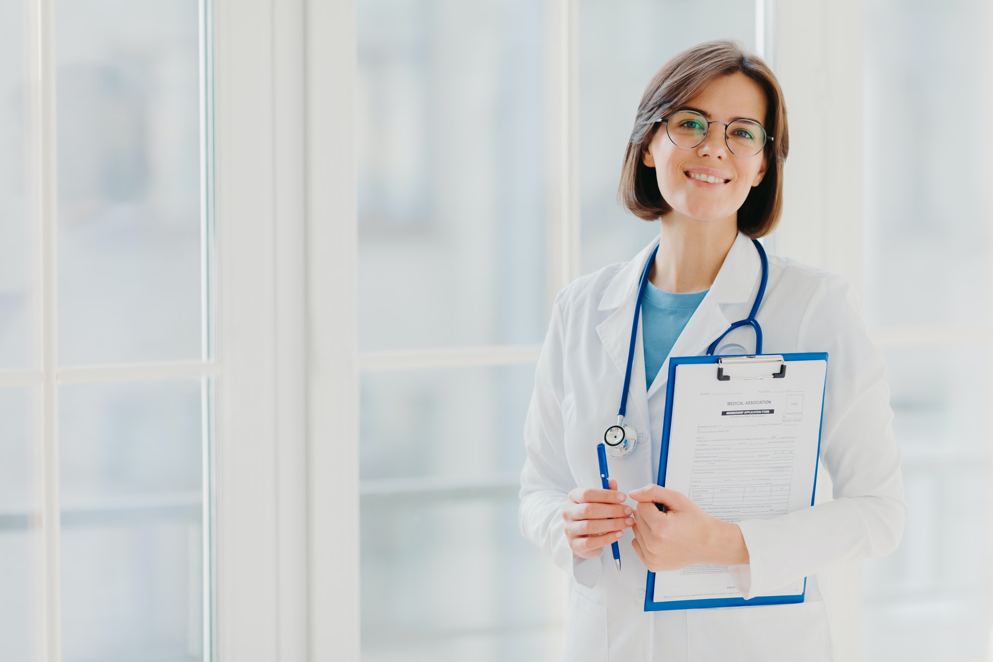 Woman doctor stands with clipboard, fills up application form, holds pen, smiles positively