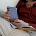 Close-up of a woman holding a smartphone and pencil, relaxing indoors with a notebook.