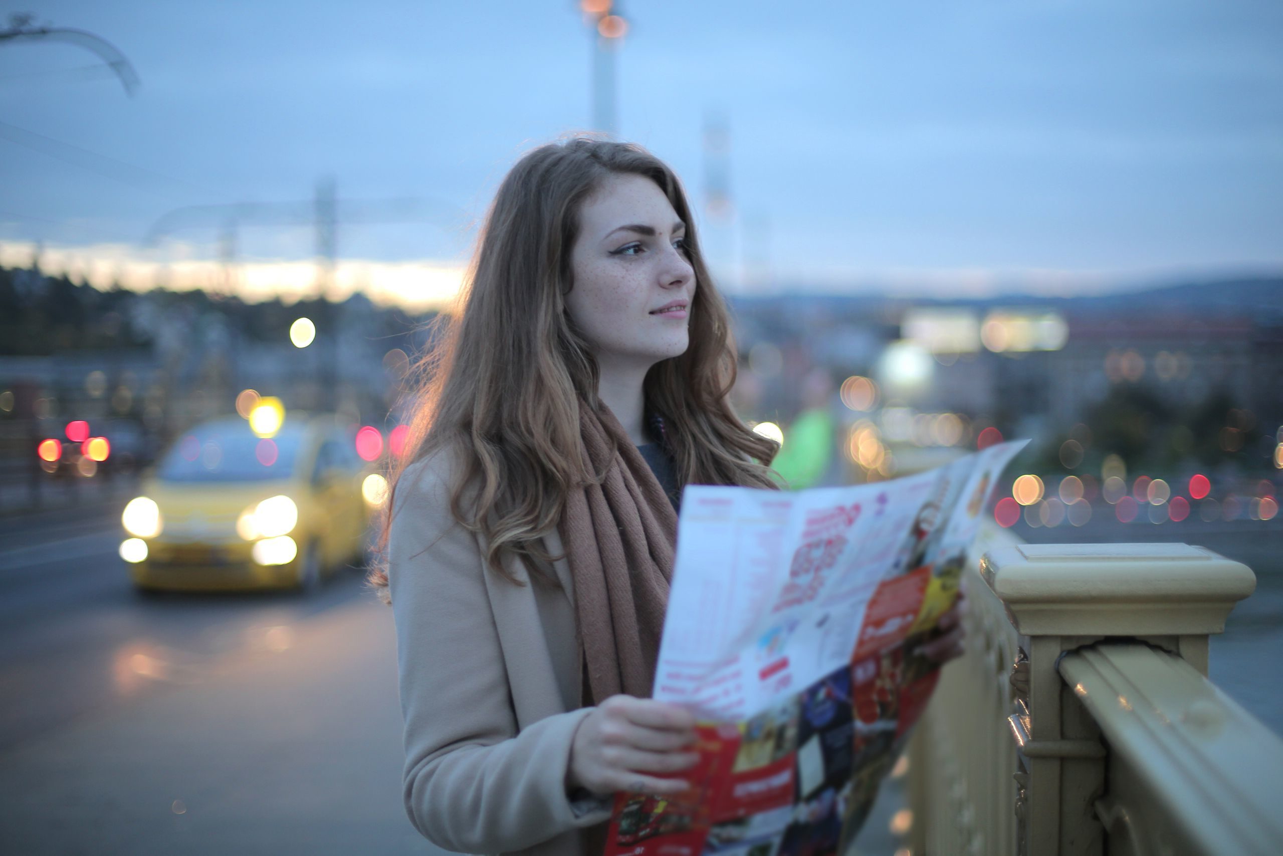 Female traveler in warm clothes standing on street with map and looking away at dusk